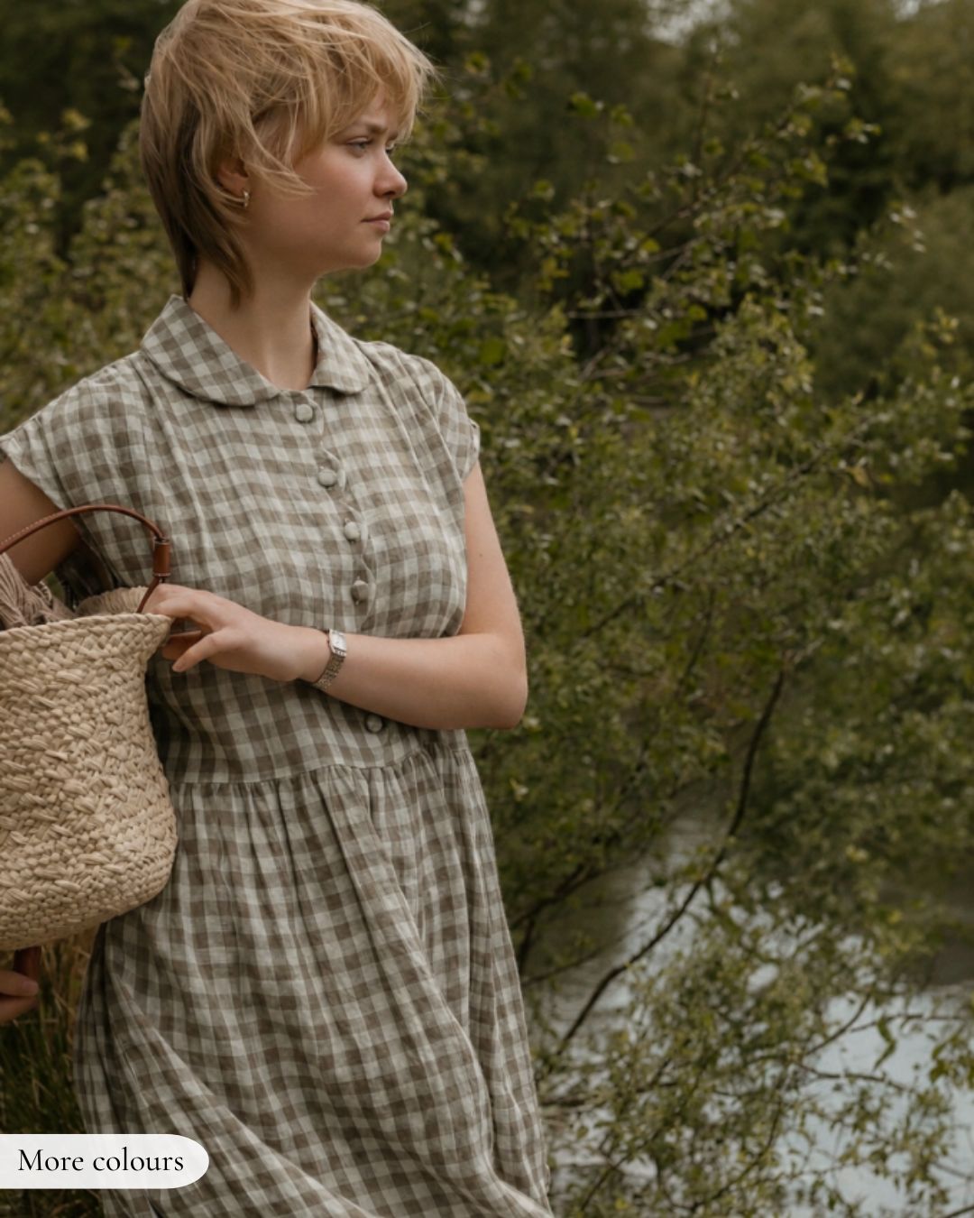 Woman in a checkered dress holding a woven bag, standing by a natural setting with trees and water.