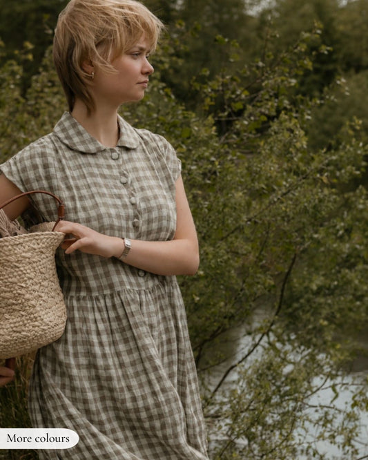 Woman in a checkered dress holding a woven bag, standing by a natural setting with trees and water.