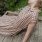 model in a brown and white checkered dress sitting on a wooden surface with greenery in the background