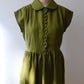 An moss green linen dress with a Peter Pan collar and scalloped button-front bodice, displayed on a vintage wooden dress form against a neutral light grey background.
