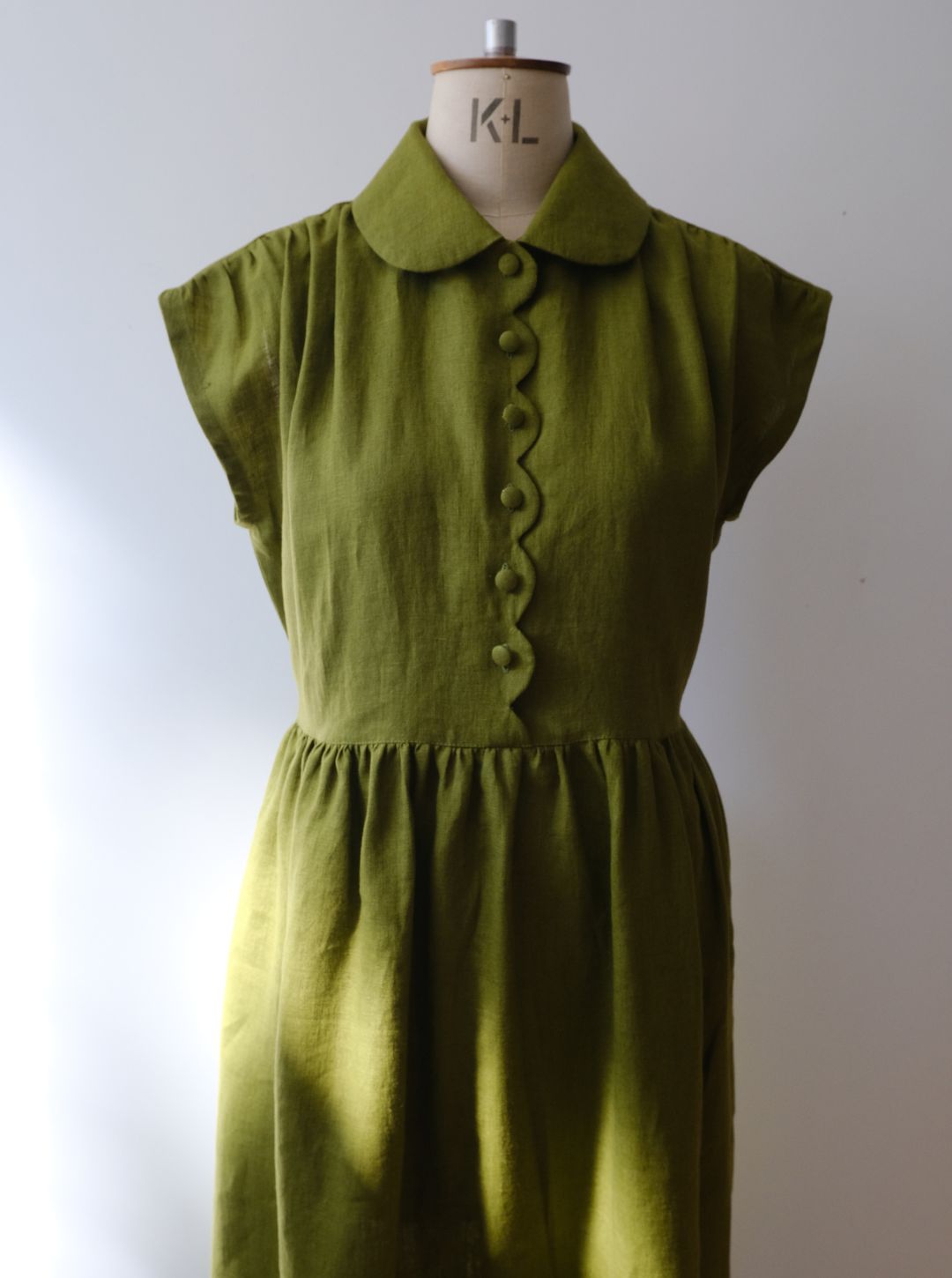 An moss green linen dress with a Peter Pan collar and scalloped button-front bodice, displayed on a vintage wooden dress form against a neutral light grey background.