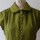 An moss green short-sleeve linen dress with a scalloped button placket and a wide Peter Pan collar, displayed on a vintage wooden dress form with the letters 'K+L' visible on the neck.