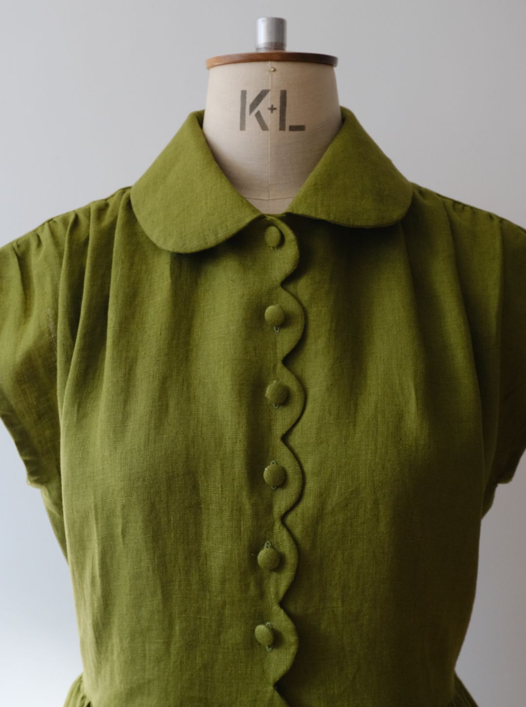 An moss green short-sleeve linen dress with a scalloped button placket and a wide Peter Pan collar, displayed on a vintage wooden dress form with the letters 'K+L' visible on the neck.