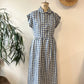 Blue and white checkered dress on a mannequin against a plain wall with a plant and straw hat in the background.