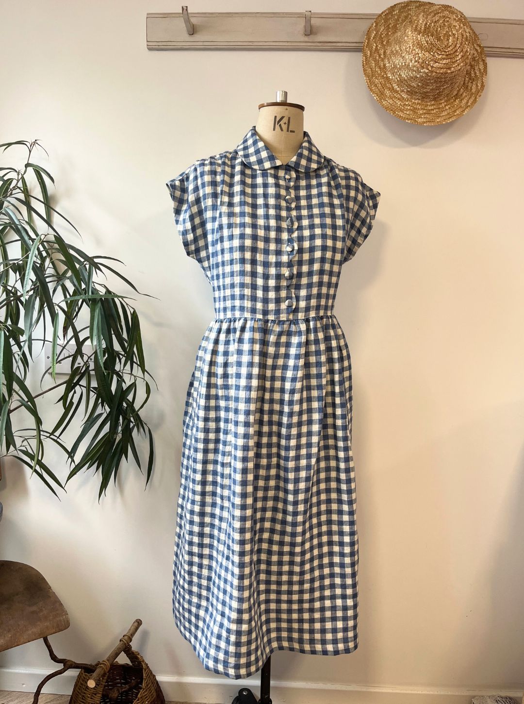 Blue and white checkered dress on a mannequin against a plain wall with a plant and straw hat in the background.