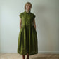 A woman stands in a room wearing a loose olive green midi dress with short cap sleeves, a buttoned collar, and a gathered waist. She is wearing black Mary Jane shoes.