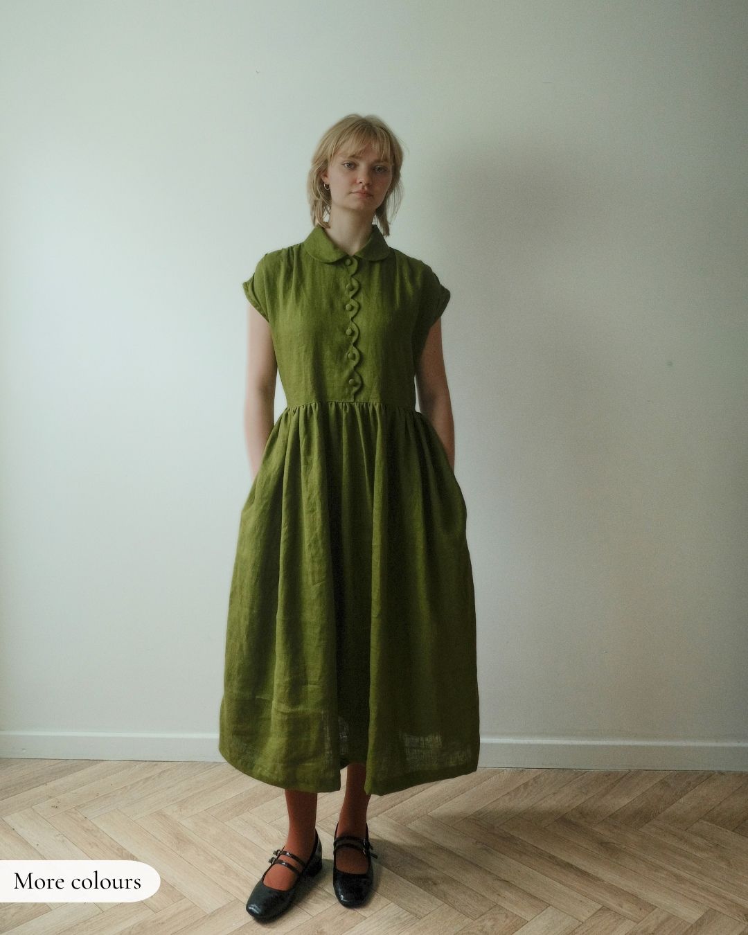 A woman stands in a room wearing a loose olive green midi dress with short cap sleeves, a buttoned collar, and a gathered waist. She is wearing black Mary Jane shoes.
