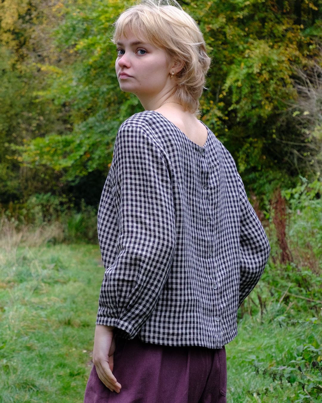 Woman wearing a black and natural linen checkered top  outdoors with greenery in the background
