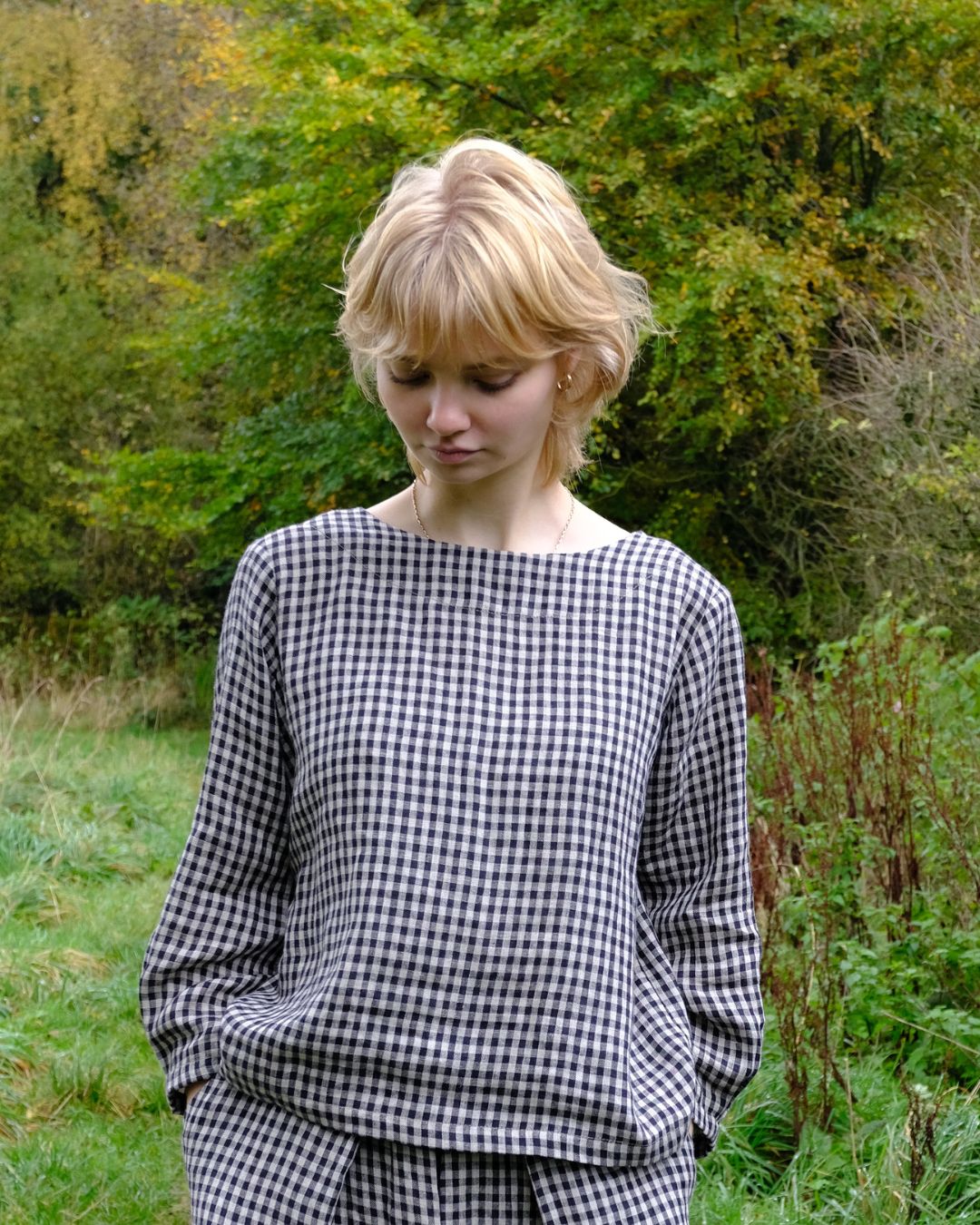 Woman wearing a black and natural checkered top standing in a natural setting with trees and grass.