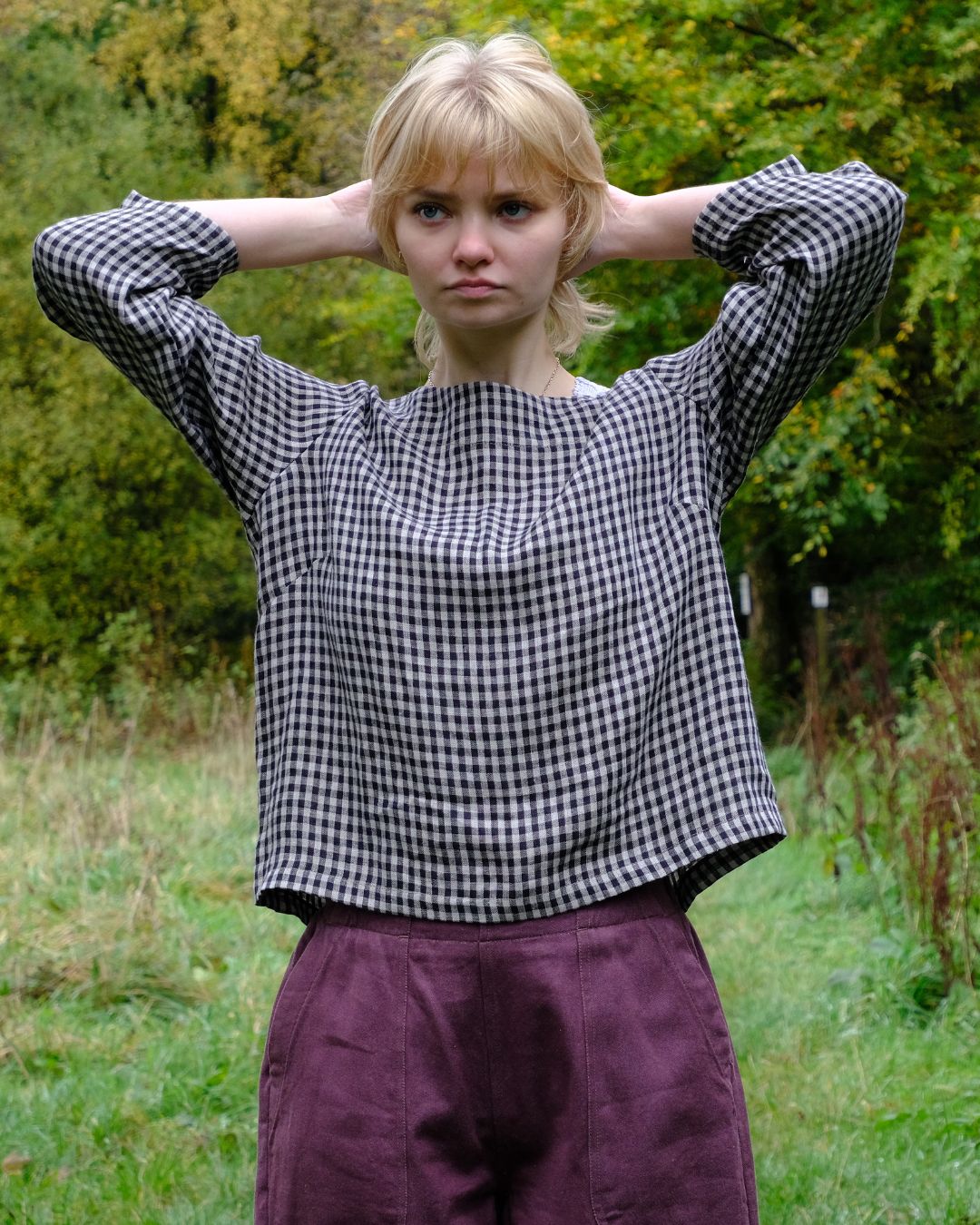 Woman wearing a black and natural checkered top and purple pants standing in a grassy area with trees.