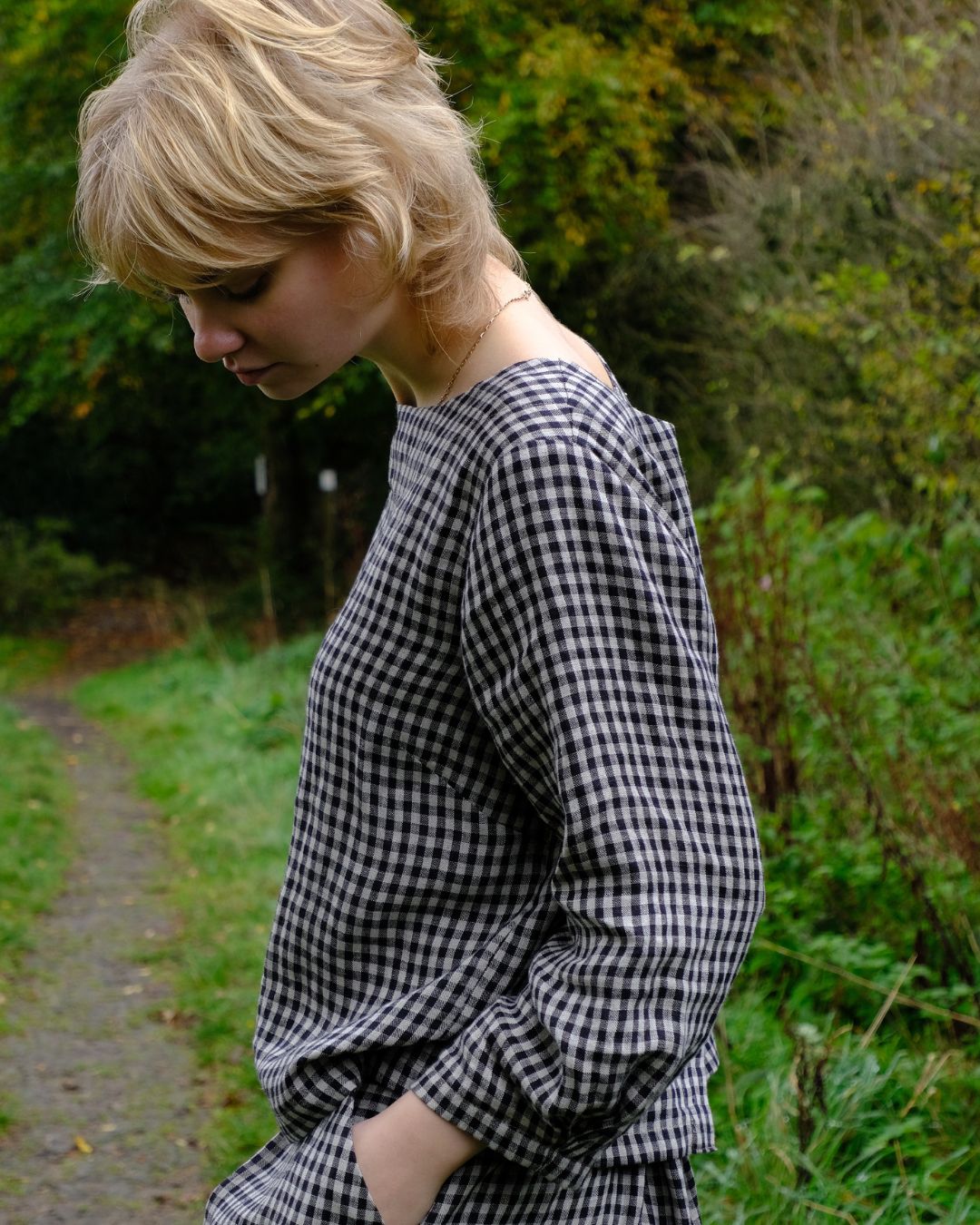 Person wearing a black and natural checkered top  standing outdoors with greenery in the background