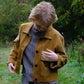 Person wearing a mustard jacket and checkered outfit standing in a grassy area with trees in the background