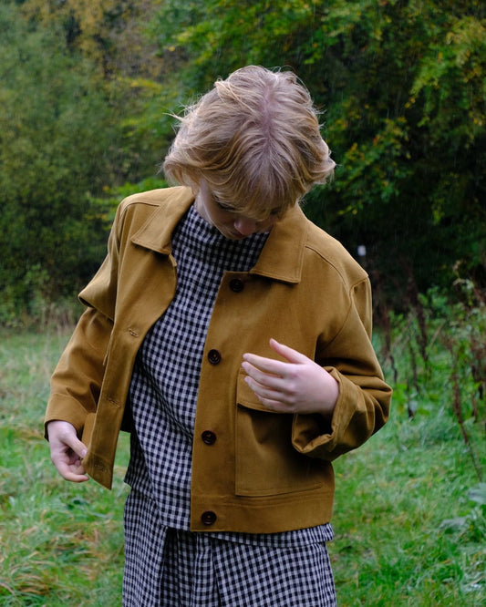 Person wearing a mustard jacket and checkered outfit standing in a grassy area with trees in the background