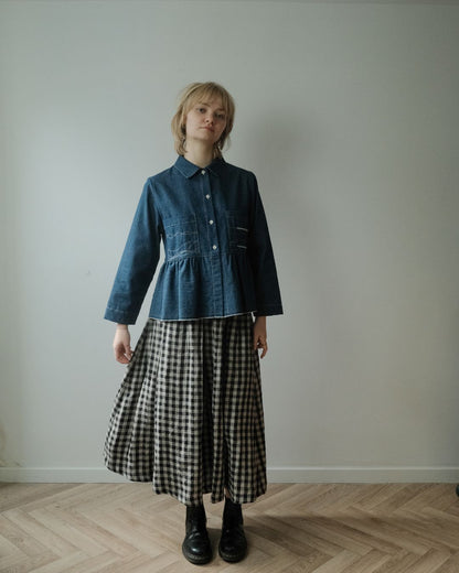 A model stands in a minimalist room with a light gray wall and wood herringbone floor, wearing a dark blue denim long-sleeve button-up shirt with a gathered waist and a black and natural  gingham pleated midi skirt.