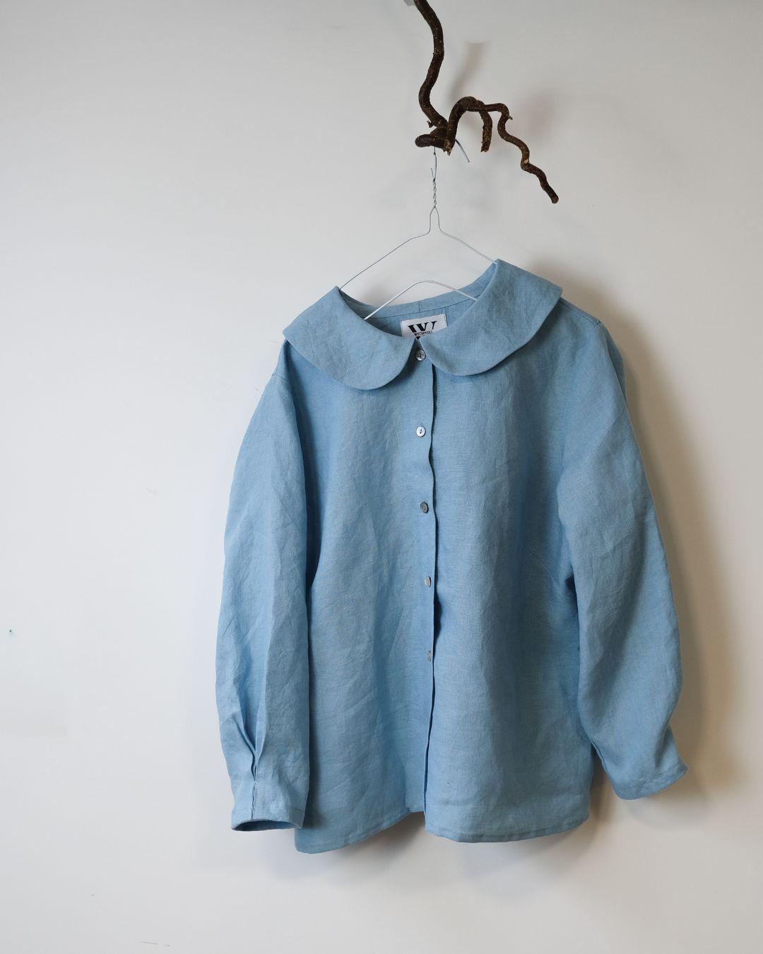 blue  linen shirt with peter pan collar and 6 buttons hanging from a twig against a white background