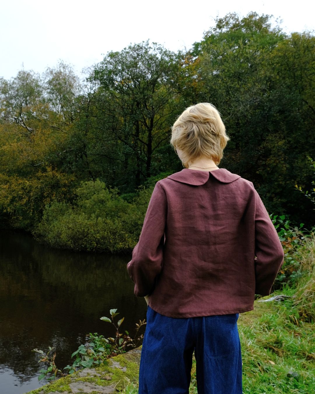 Person wearing a maroon jacket and blue pants standing by a body of water with trees in the background