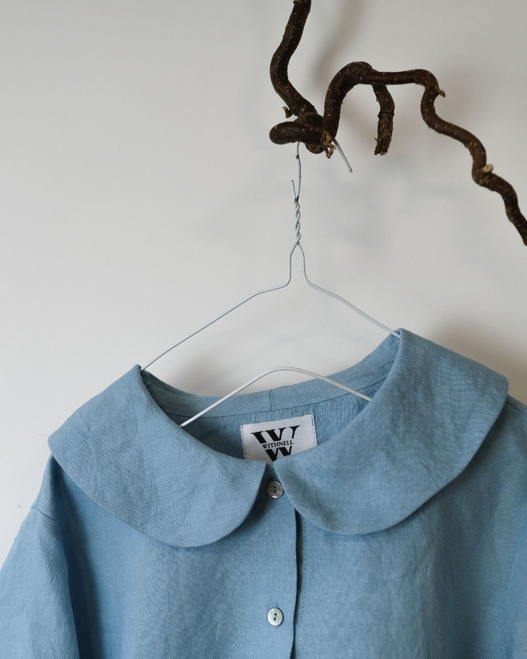 blue  linen shirt with peter pan collar and 6 buttons hanging from a twig against a white background