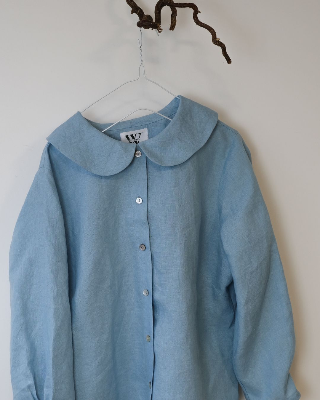 blue  linen shirt with peter pan collar and 6 buttons hanging from a twig against a white background