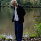 A person standing outdoors by a lake, wearing a dark jacket over a white shirt, loose dark blue trousers, and black shoes. Trees and foliage are visible in the background.