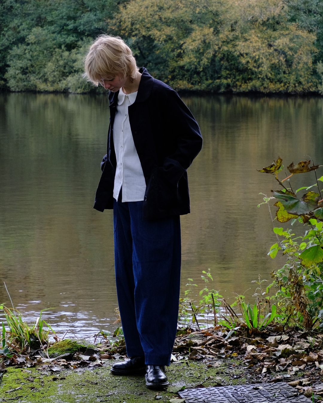 A person standing outdoors by a lake, wearing a dark jacket over a white shirt, loose dark blue trousers, and black shoes. Trees and foliage are visible in the background.