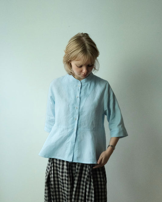 A young girl with blonde hair looking down, wearing a light blue linen button-up shirt with three-quarter sleeves and a black-and-white gingham skirt, standing against a plain light gray wall.