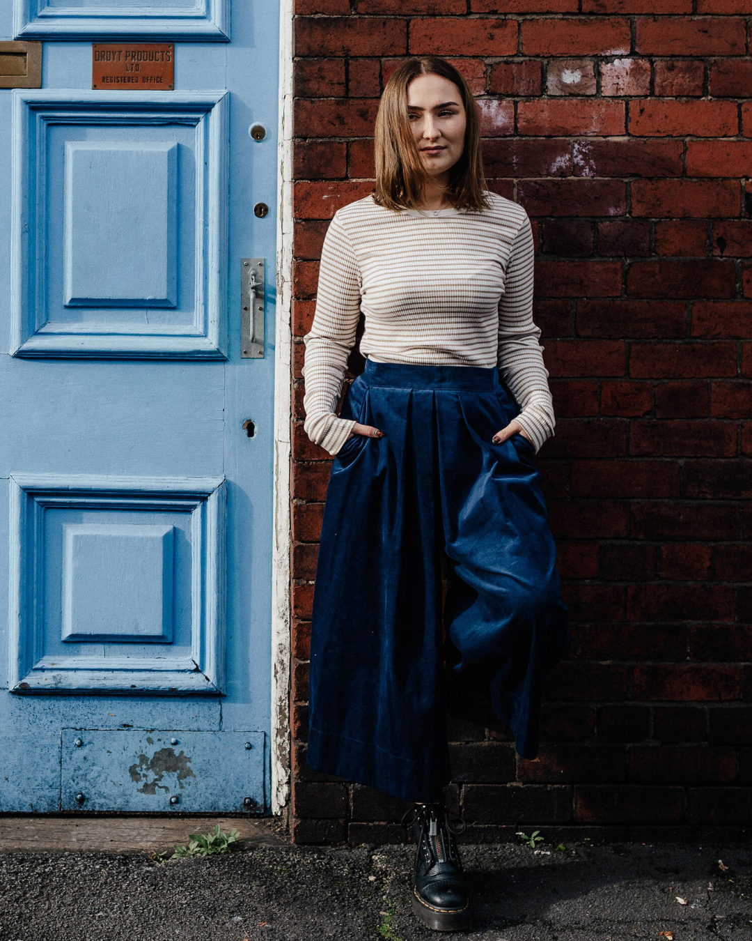 Woman standing by the side of a blue door wearing stripe top and blue culottes