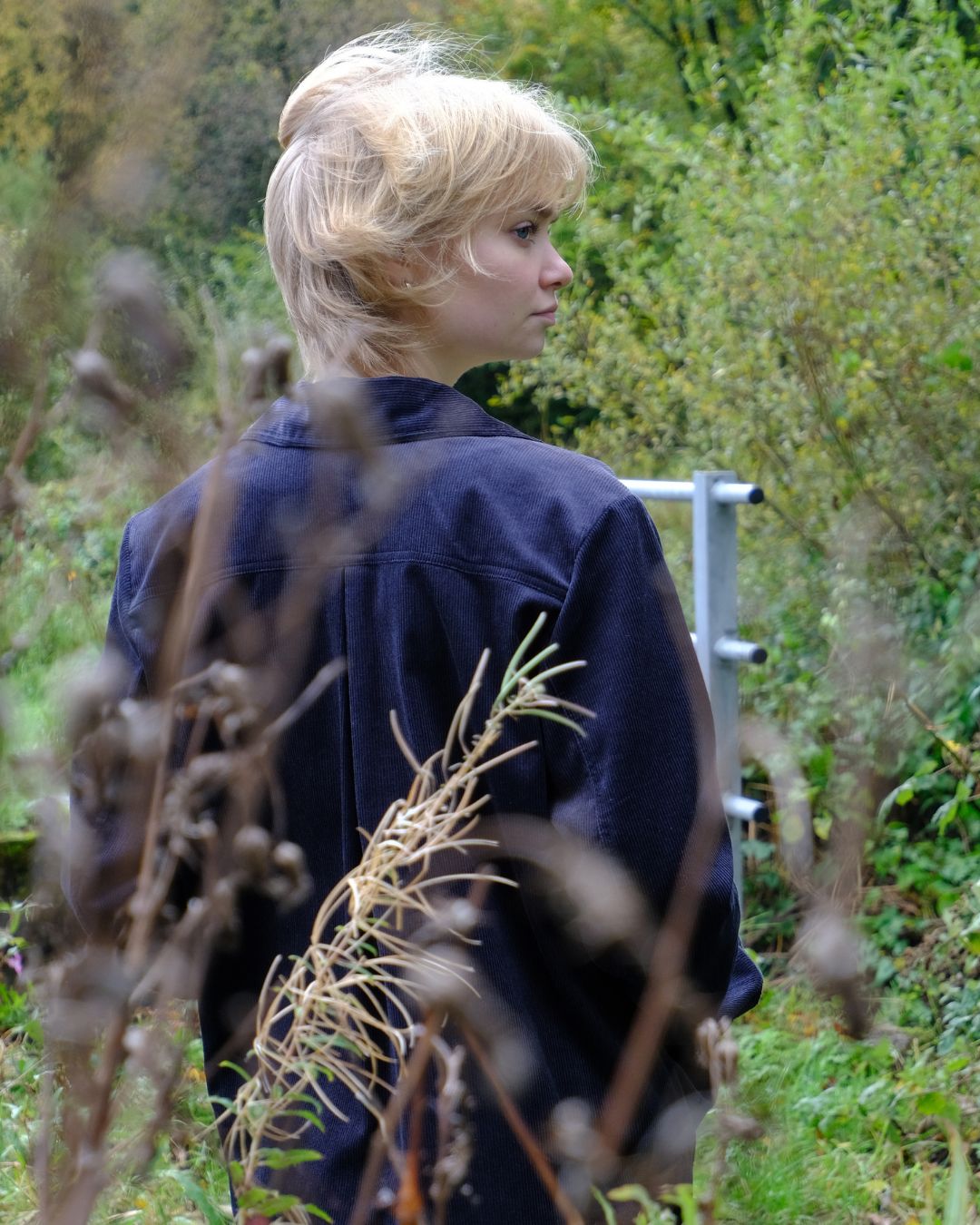 Person with blonde hair standing in a natural setting with greenery and a metal structure.