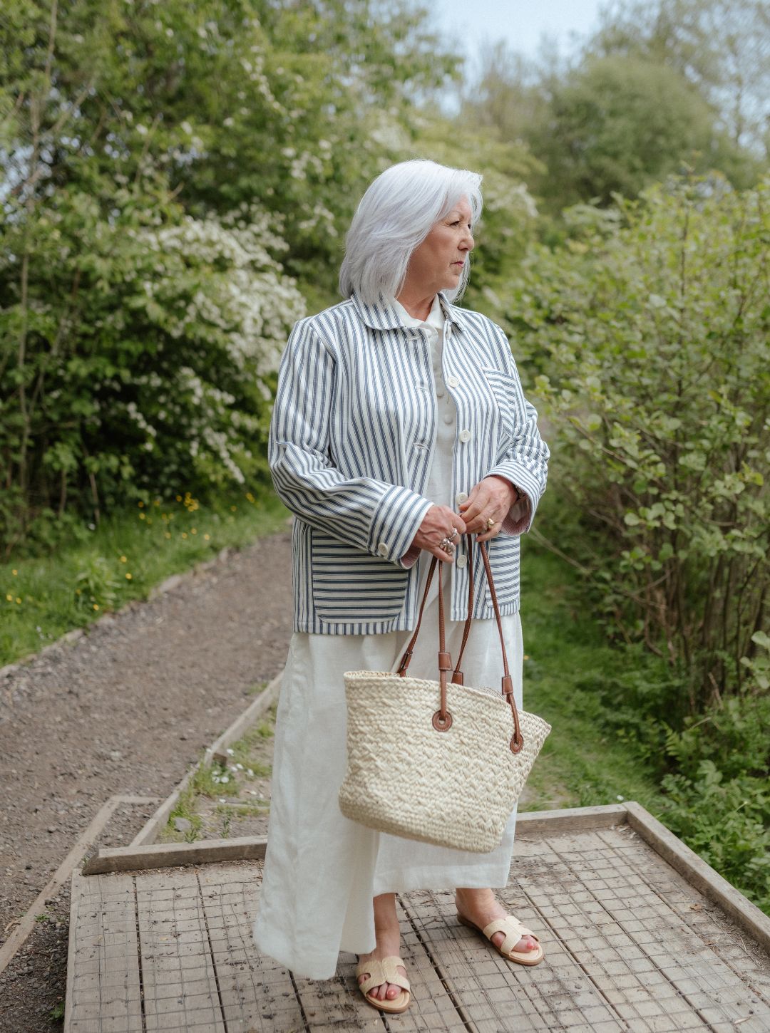 Woman standing outdoors holding a woven bag, surrounded by greenery