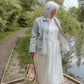 Woman in a white dress and striped jacket standing by a path with greenery and water in the background