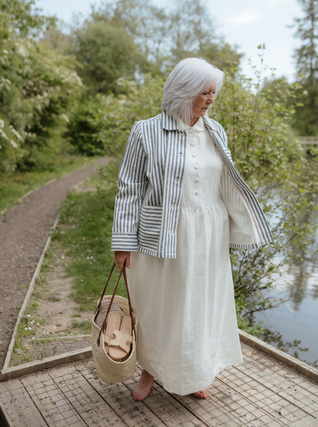 Woman in a white dress and striped jacket standing by a path with greenery and water in the background