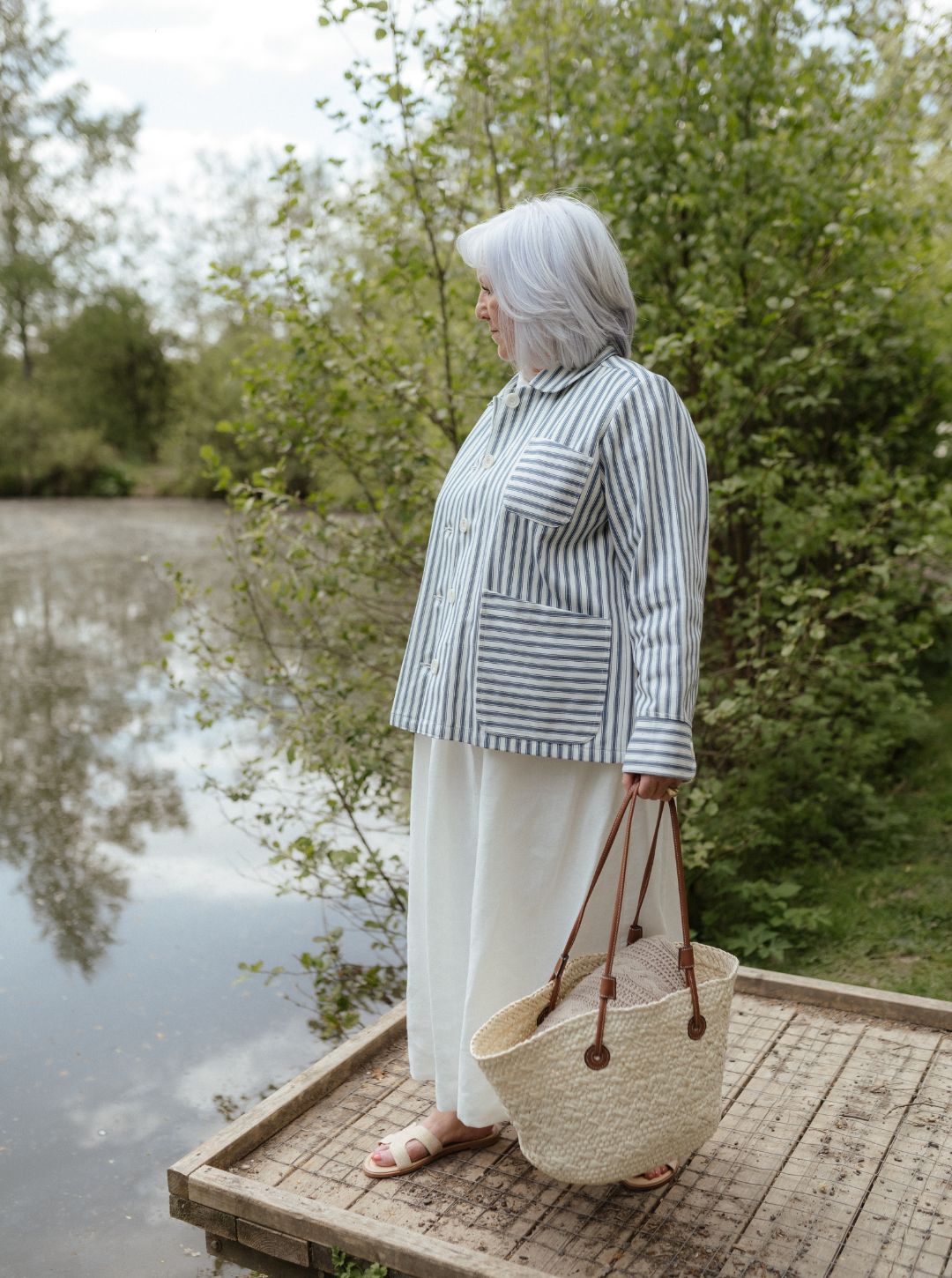 Woman standing on a wooden platform by a body of water, holding a woven bag.
