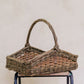 A rectangular wicker trug style basket with a handle, displayed on a cylindrical support.