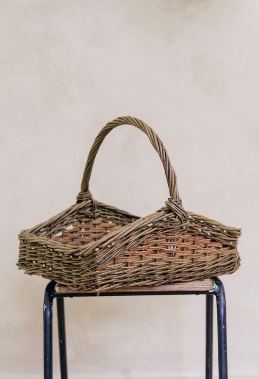 A rectangular wicker trug style basket with a handle, displayed on a cylindrical support.