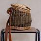 A hand woven basket made of willows featuring a leather strap and wooden fastener, displayed on a wooden stool.