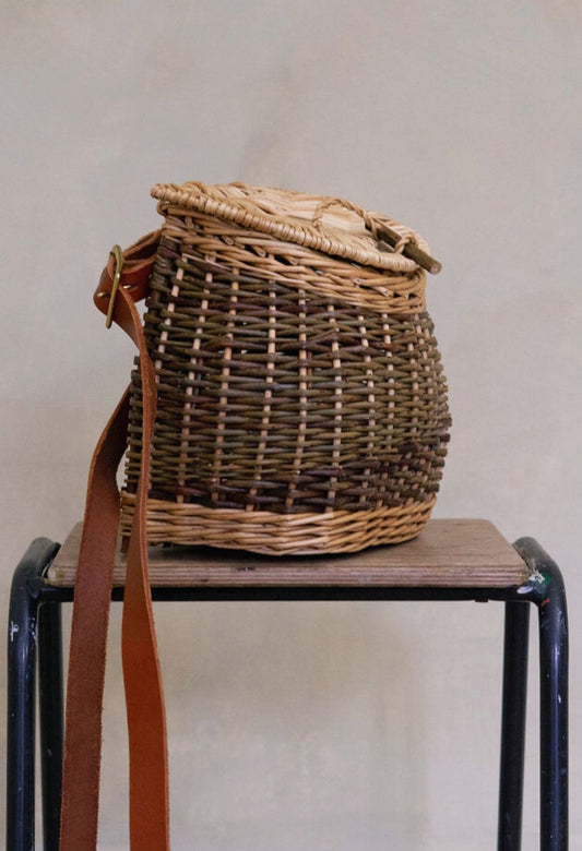 A hand woven basket made of willows featuring a leather strap and wooden fastener, displayed on a wooden stool.