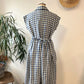 Blue and white checkered dress on a mannequin against a white wall with a plant and hat in the background.
