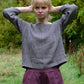 Woman wearing a black and natural checkered top and purple pants standing in a grassy area with trees.