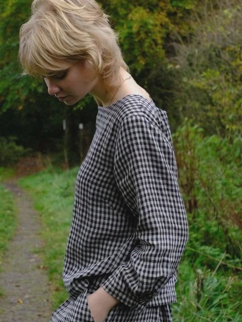 Woman wearing a black and natural checkered top and standing in a grassy area with trees.